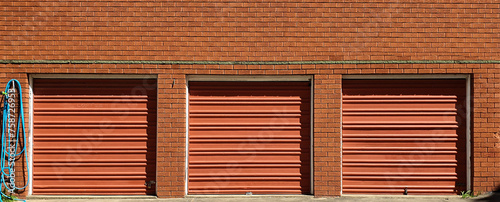 three single brown colored garage doors in a row against red brick building with blue hose hanging on side of building
