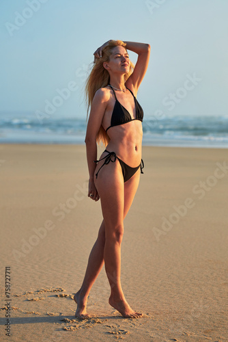 A slender woman in a swimsuit walks along the seashore