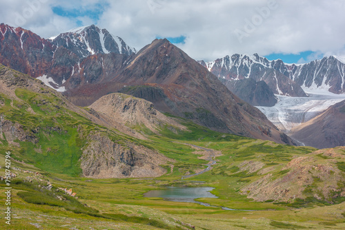 Vivid mountain scenery with beautiful lake among green hills and rocks in alpine valley with view to large snowy rocky peaked top, sharp rockies of red color and big glacier under clouds in blue sky.