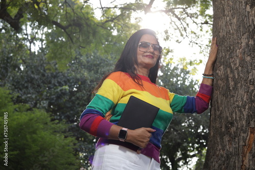 Portrait of mid aged woman holding a black color almanac in hand, sunlight, green park serene environment. lady leaning against tree.