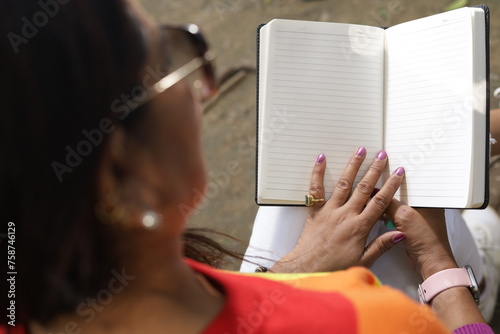 Portrait of mid aged woman holding a black color almanac in hand, reading personal almanac. sunlight, green park environment. book promotion, blank pages. photo