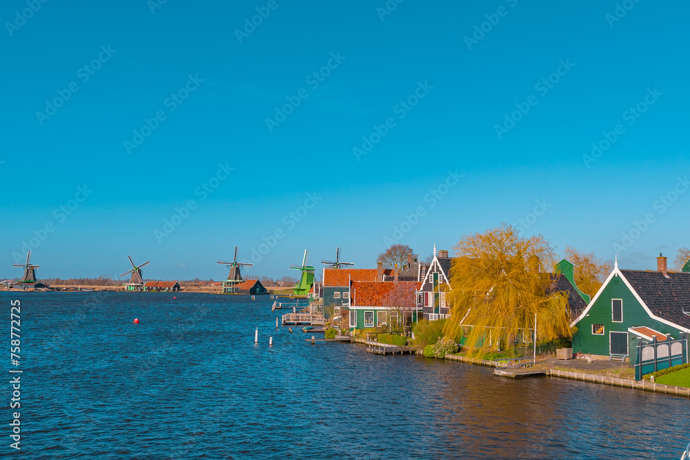 Naklejka premium Beautiful Dutch scenery panorama of Zaanse Schans village in Netherlands in spring time. Ancient windmills at the water's edge in the background with the blue sky in sunny weather