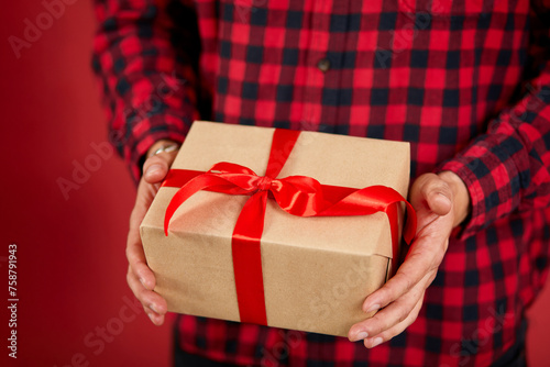 Fototapeta Naklejka Na Ścianę i Meble -  Close-up of hands holding a gift box with a red ribbon against a red background.