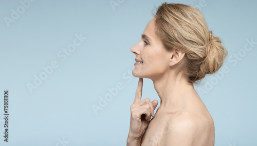 Mature woman pointing finger at toned skin on her chin, thanks to anti-aging cosmetics and face contouring with beauty injections, side view on blue background