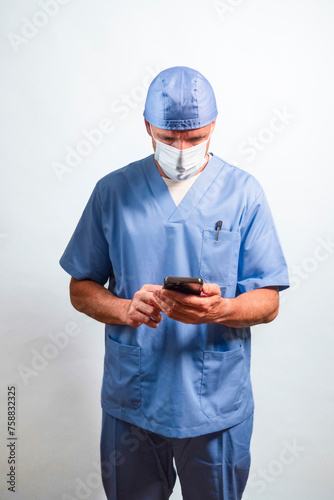 Portrait of a doctor in light blue gown, mask and cap using a smartphone.