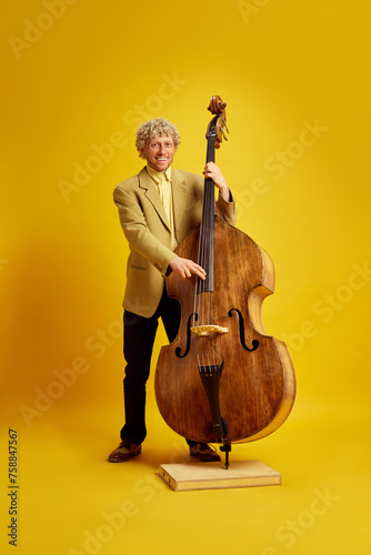 Stylish man in suit paying double bass, making classical music live performance against bright yellow background. Concept of music, performance, art, entertainment, festival, performance, ad