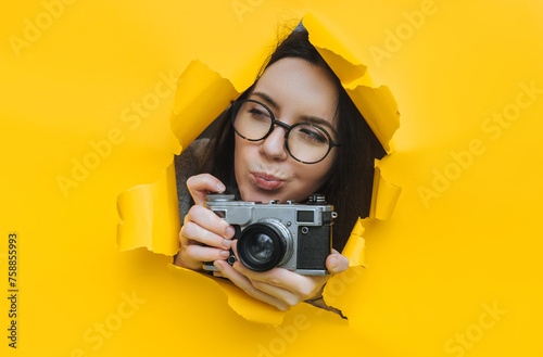 A paparazzi girl in glasses with a rare SLR camera looks out from her hiding place and carefully looks at what is happening. Yellow paper, torn hole. Tabloid press.Looking for a story for stock photos