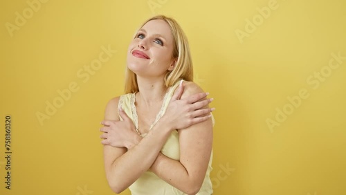 Beaming blonde beauty, young woman hugs herself in pleasure, wearing sleeveless t-shirt over yellow background. portrait of confidence, self love, and happiness.