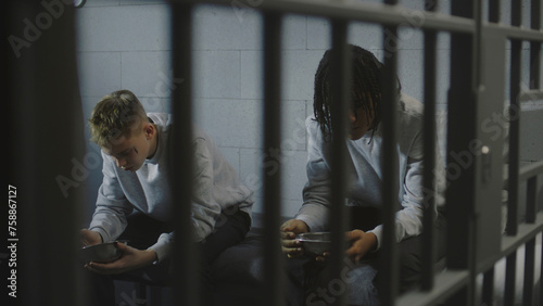 Schilderij op canvas Two multiethnic teenage prisoners sit on bed in prison cell and eat food from iron bowls