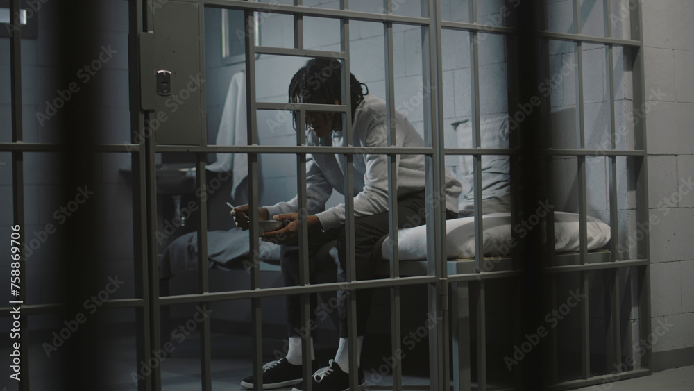 African American young prisoner eats disgusting prison food from iron bowl sitting on bed in prison cell. Teenage criminal, inmate serves imprisonment term for crime in jail. Youth detention center. - obrazy, fototapety, plakaty 