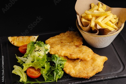 Crispy golden chicken schnitze served with fresh salad and french fries on a black stone plate. photo