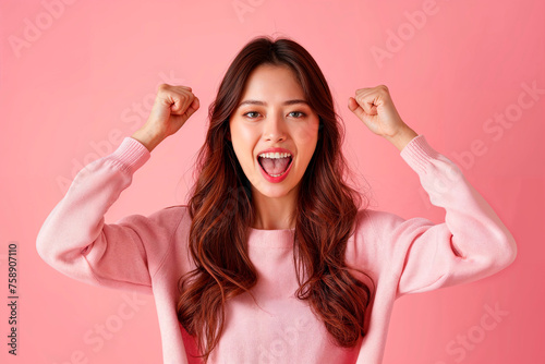 una mujer con los brazos en alto y los puños en alto, con un fondo rosa, hace una mueca graciosa