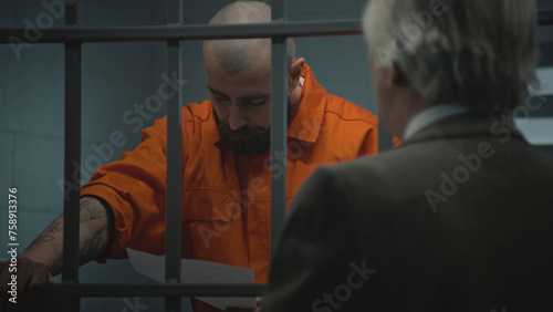Fotografía Prisoner in orange uniform leans on prison cell bars, talks with advocate and reads lawyer contract