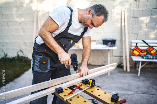 Mature male carpenter using electric drill on a wooden plank outdoors