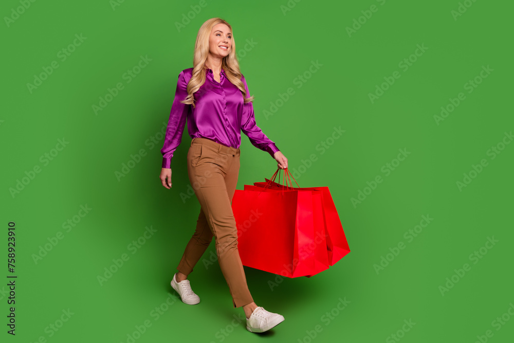 Full body profile photo of lovely positive lady hold mall bags look walk empty space isolated on green color background Full body profile photo of lovely positive lady hold mall bags look walk empty space isolated on green color background