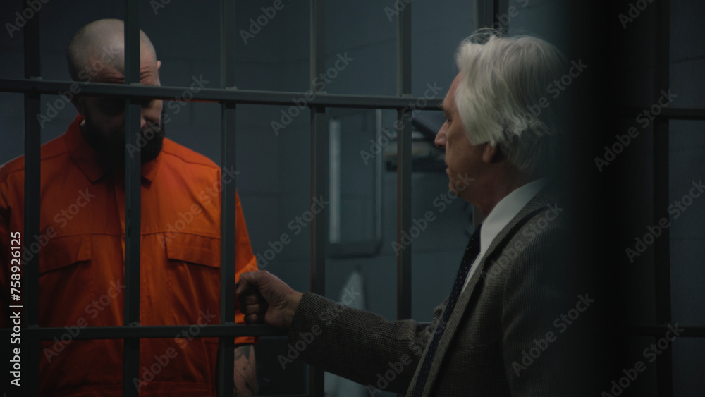 Inmate in orange uniform stands behind bars in prison cell, talks with advocate, reads lawyer contract. Gangster serves imprisonment term for crime in jail. Detention center or correctional facility.