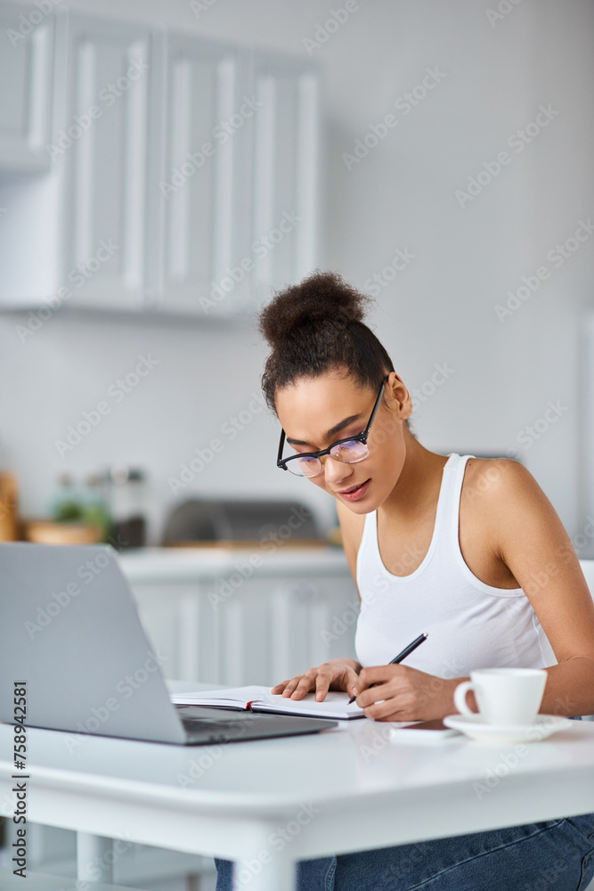 curly african american woman in glasses working from home remotely on her laptop, taking notes curly african american woman in glasses working from home remotely on her laptop, taking notes