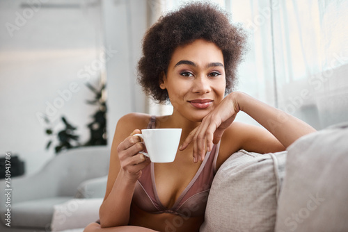 happy african american woman in lingerie holding cup of coffee and looking at camera