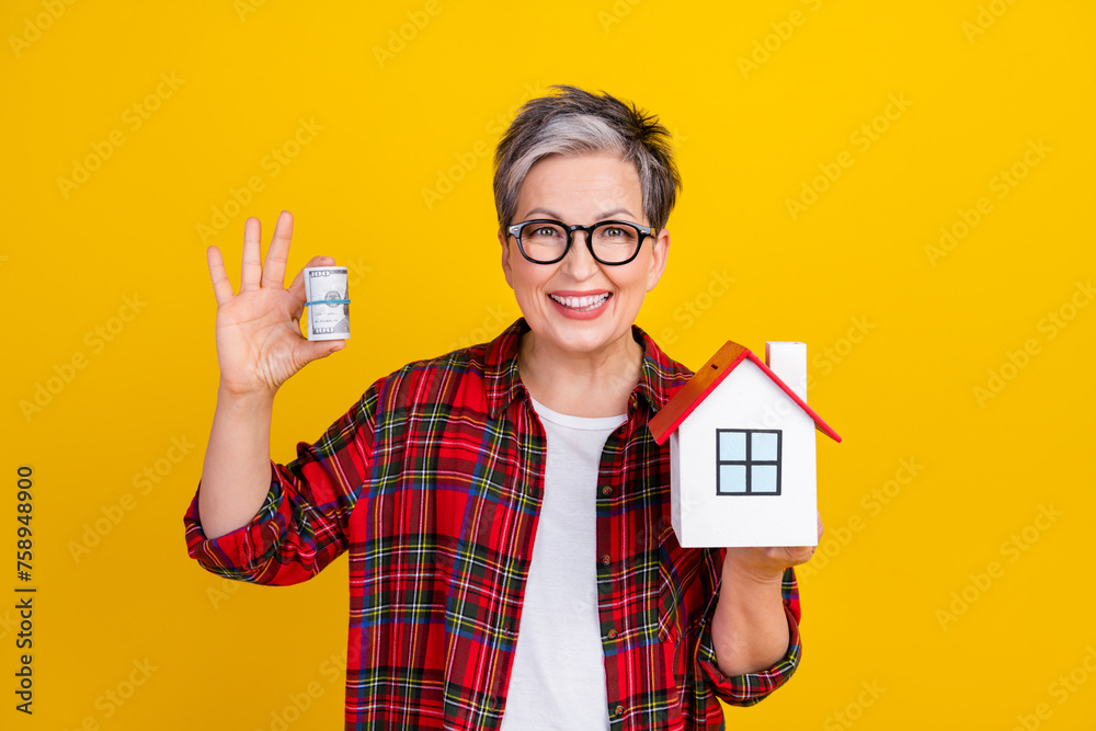 Photo of cheerful grandmother hold rolled stack dollars little house isolated on yellow color background - obrazy, fototapety, plakaty 
