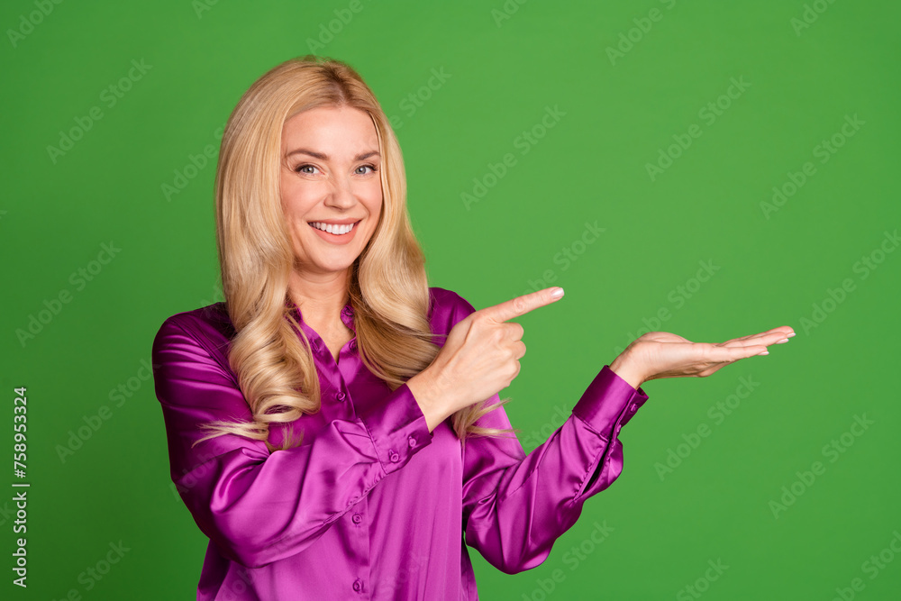 Photo of stunning cheerful lady toothy smile indicate finger arm hold empty space ad isolated on green color background Photo of stunning cheerful lady toothy smile indicate finger arm hold empty space ad isolated on green color background