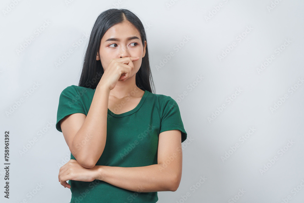 Young Asian frustrated woman with green t-shirt pinches her nose with disgust on her face, reacting to a bad smell in isolation over background. - obrazy, fototapety, plakaty 