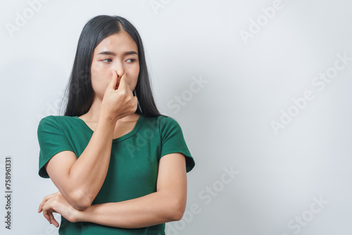 Young Asian frustrated woman with green t-shirt pinches her nose with disgust on her face, reacting to a bad smell in isolation over background.