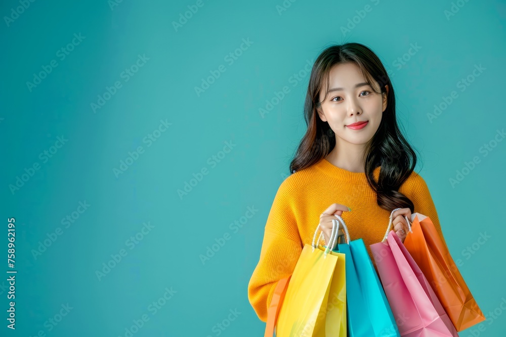 Young Asian Woman in Orange Sweater Smiling with Colorful Shopping Bags on Blue Background Young Asian Woman in Orange Sweater Smiling with Colorful Shopping Bags on Blue Background