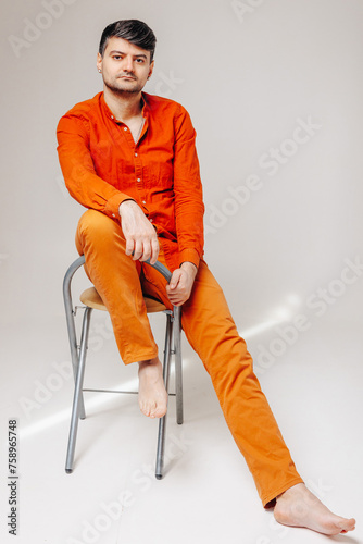 Man in bright orange clothes sits on a chair with his leg raised and looks into the camera lens