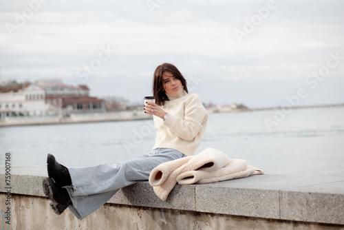 A girl with a stylish bob haircut is walking around the city, drinking coffee against the background of water and buildings. The woman is wearing a white faux fur sheepskin coat and jeans