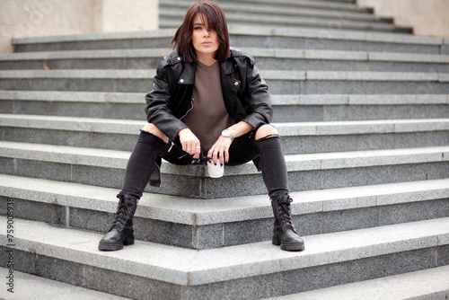 A beautiful girl with a stylish bob haircut is walking around the city, drinking coffee or tea, sitting on the stairs. The woman is wearing a black leather, sweater, torn jeans and boots