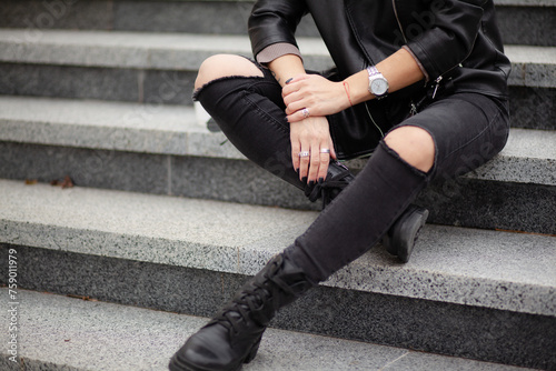 A beautiful girl with a stylish bob haircut is walking around the city, sitting on the stairs. The woman is wearing a black leather, sweater, torn jeans and boots