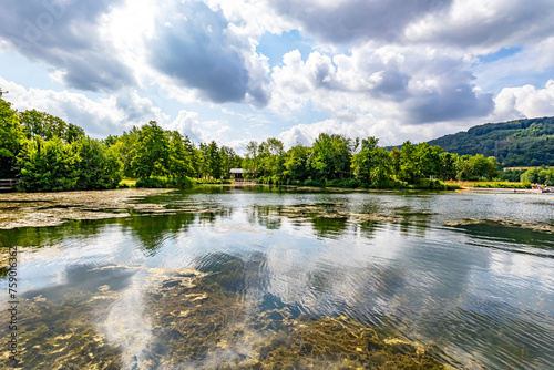Fototapeta Naklejka Na Ścianę i Meble -  Echternach lake in panoramic nature landscape, algae and reflections on water surface, green leafy trees against blue sky with white clouds in background, sunny summer day in Luxembourg