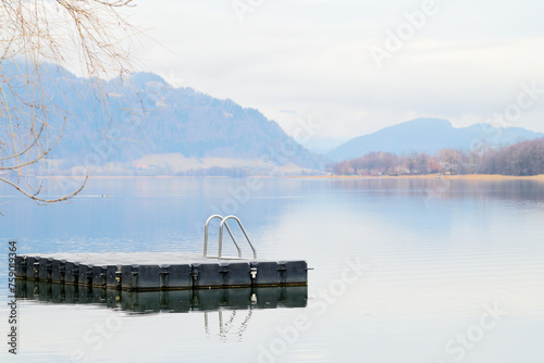 Spokój nad górskim jeziorem zimą. Drewniana platforma ze srebrną drabinką dla pływaków i plażowiczów. W tle zamglone góry. © Tomasz