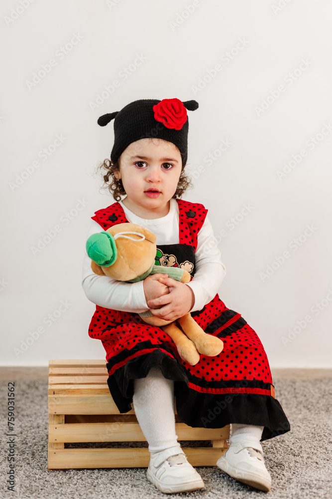 Cute little girl on wooden box dressed in a vivid red gown, embracing a soft toy in the spirit of Easter
