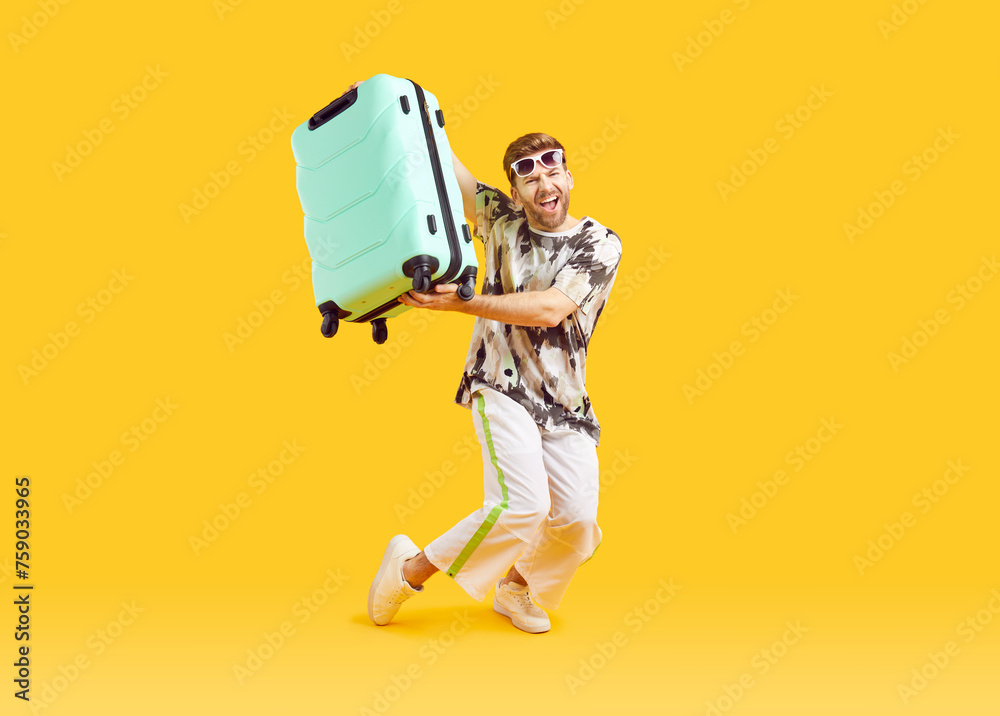 Full body photo of a funny young happy man in sunglasses carrying his blue suitcase is going on summer holiday trip and having fun on a yellow background. Vacation and travel concept. - obrazy, fototapety, plakaty 