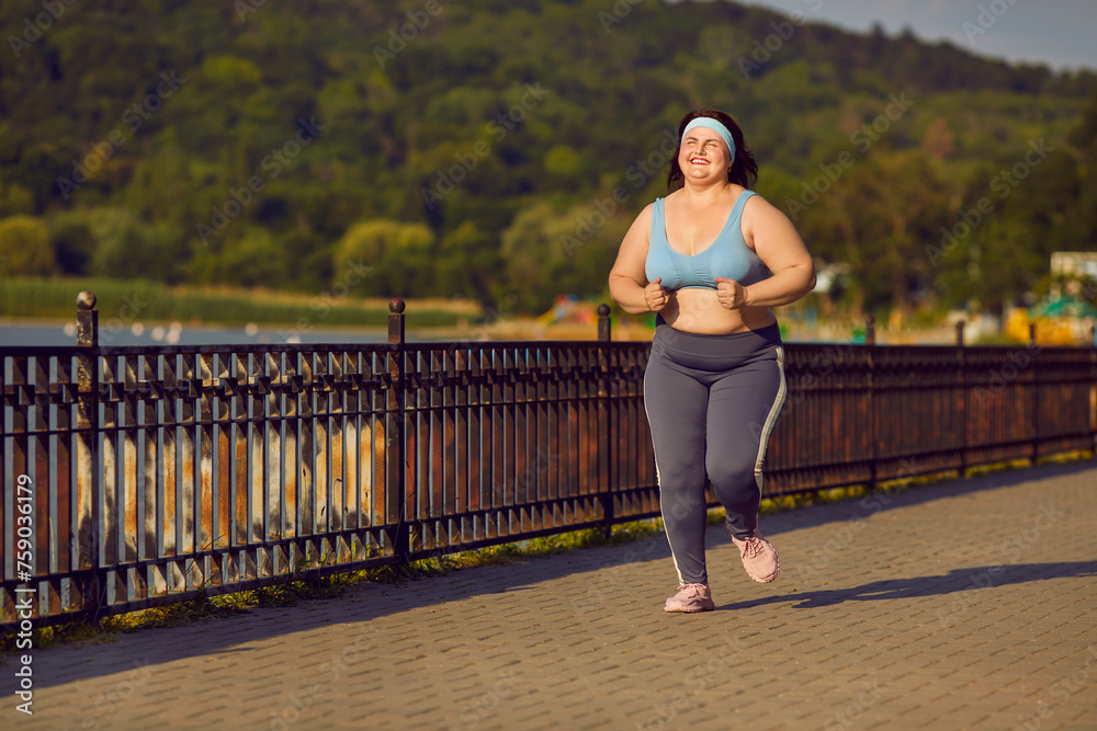 Overweight happy smiling fat woman running in the summer park across the bridge. Plus size girl wearing sportswear jogging outdoors. Weight loss, body positive, sport and fitness lifestyle concept.