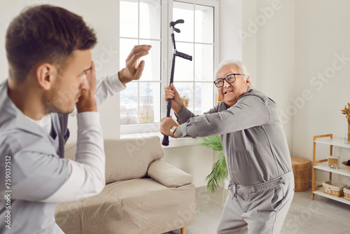 Angry elderly senior man going to hit young male caregiver or nurse with his crutch. Enraged exasperated male pensioner standing at home and threatening scared medical caretaker with cane. photo