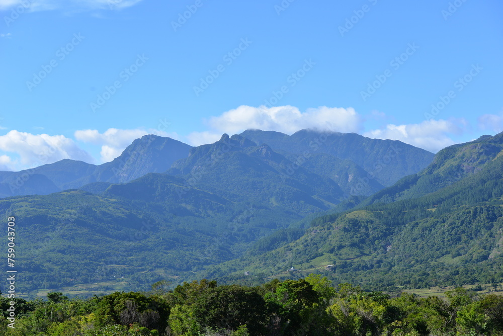 Before us unfolds the heart of nature's realm, where mountain shadows and valleys bask under a sky of boundless blue, whispering ancient tales of the earth's splendor.