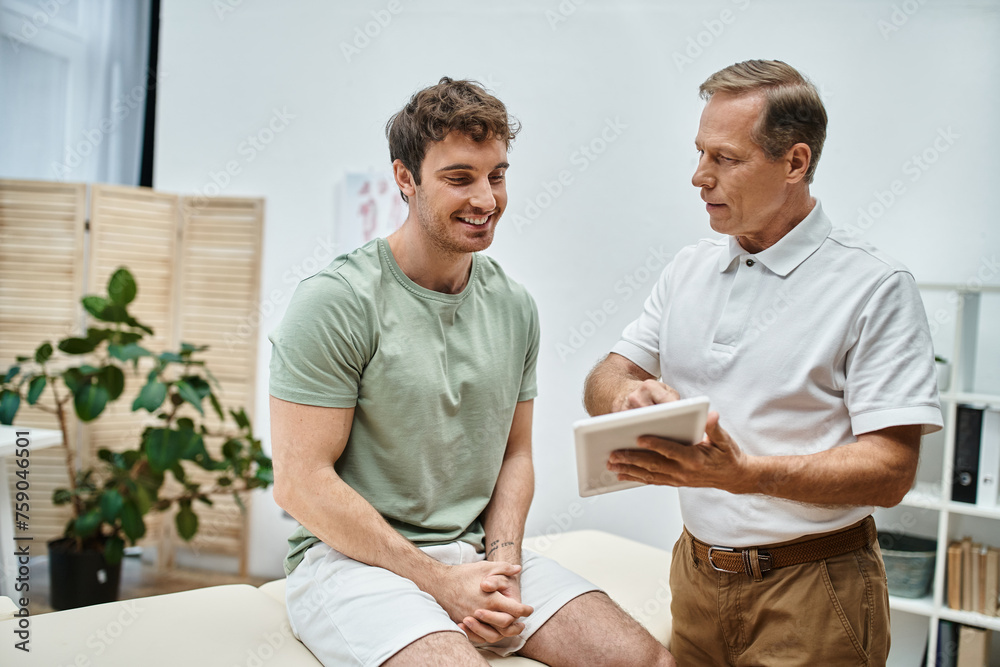 good looking jolly doctor in uniform holding tablet and talking to his joyous patient on appointment good looking jolly doctor in uniform holding tablet and talking to his joyous patient on appointment