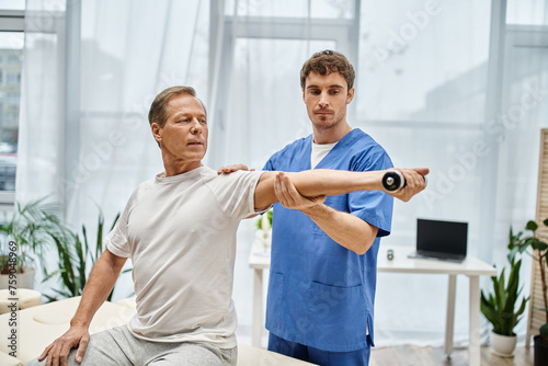 handsome doctor in blue robe helping his mature patient in casual attire to use dumbbell in hospital