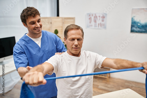 joyous attractive doctor in blue robe helping his mature patient to use resistance band in hospital