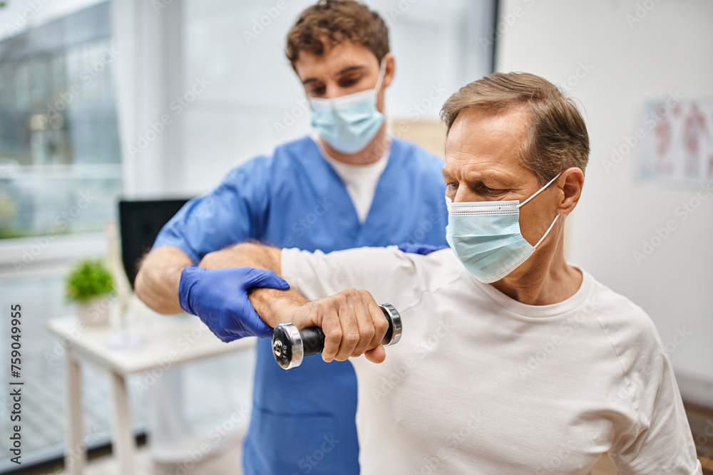 devoted handsome doctor with mask and gloves helping his patient to use dumbbells on appointment