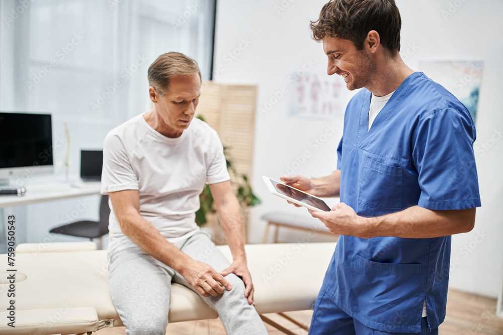 Fototapeta premium cheerful handsome doctor in blue robe holding tablet and listening to his mature patient in hospital