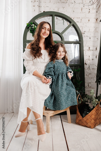 Mother's day. Elegant young mom and little cute daughter sit and smile, embrace on wooden chair at living room at home, look at camera. Trust, support and love between mommy and child. Children day