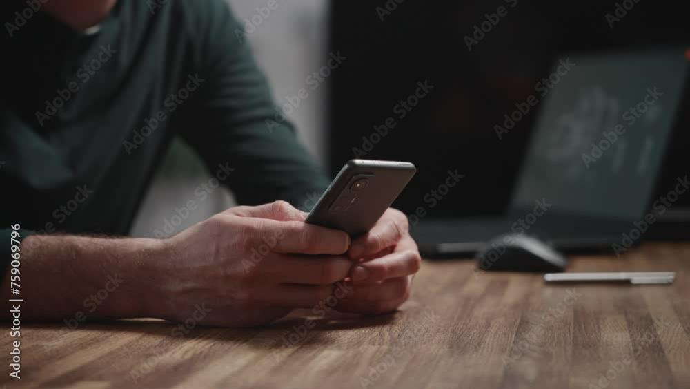 Close-up of male hands using a modern smartphone. A man is sitting at a table and using online apps. A laptop is in the background. Close-up of male hands using a modern smartphone. A man is sitting at a table and using online apps. A laptop is in the background.