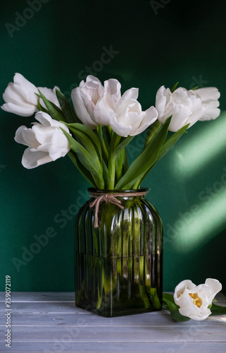 Bouquet of white tulips in green glass vase on green background with sunlight. Vase of flowers stands on wooden table. Concept: greeting card, spring holidays. Copy space. Verticale