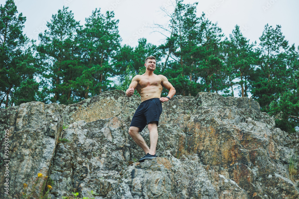 A muscular young man sits on the rocks near the forest. Relief and muscular physique of a man.