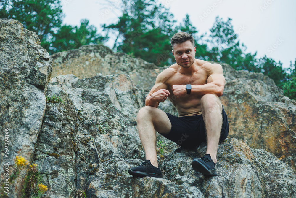 A muscular young man sits on the rocks near the forest. Relief and muscular physique of a man. A muscular young man sits on the rocks near the forest. Relief and muscular physique of a man.