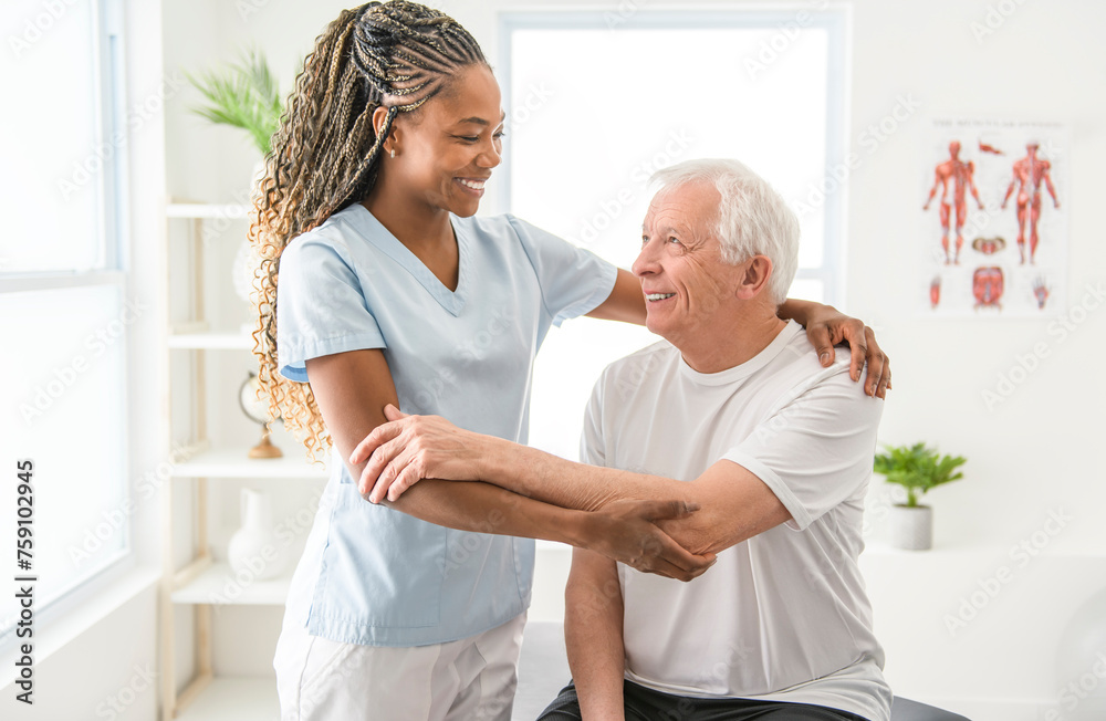 Fototapeta premium Black physiotherapist helping senior man with in clinic. Elderly man undergoing physiotherapy treatment for injury