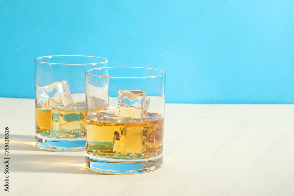 Whiskey with ice cubes in glasses on white table against light blue background, space for text Whiskey with ice cubes in glasses on white table against light blue background, space for text